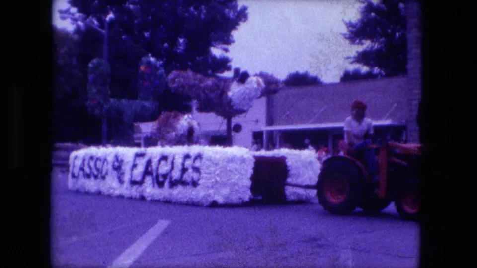 The EAGLES float passes along a Fort Wayne street behind a red tractor, with greenery and low buildings in the background.