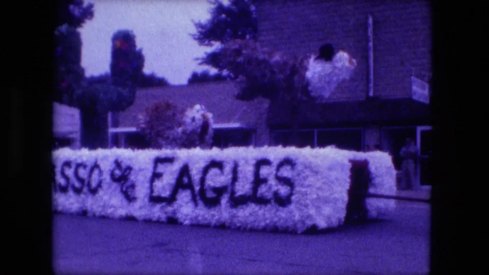 The white floral EAGLES float continues past the camera, with greenery on top and people partly visible near the right side.