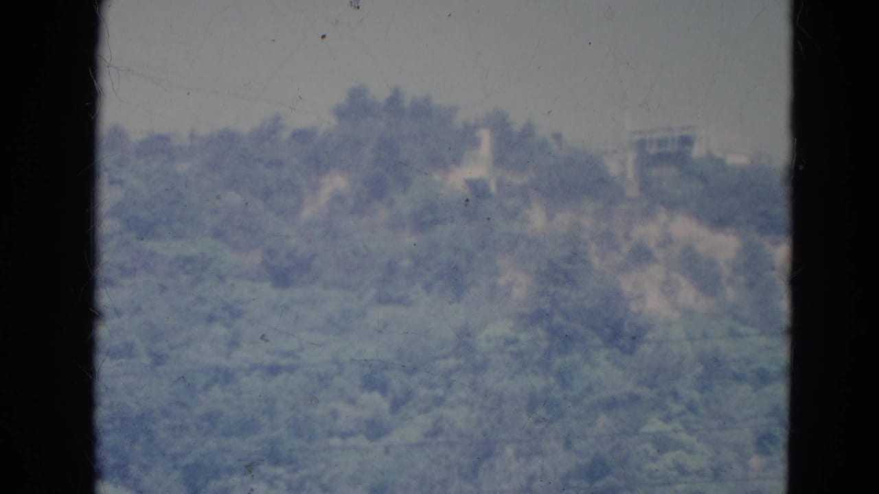 1976 Minnesota 8mm frame showing a hazy, tree-covered ridge with faint structures, seen through a dark window frame.