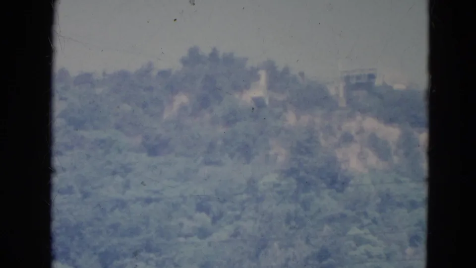 1976 Minnesota 8mm frame showing a hazy, tree-covered ridge with faint structures, seen through a dark window frame.