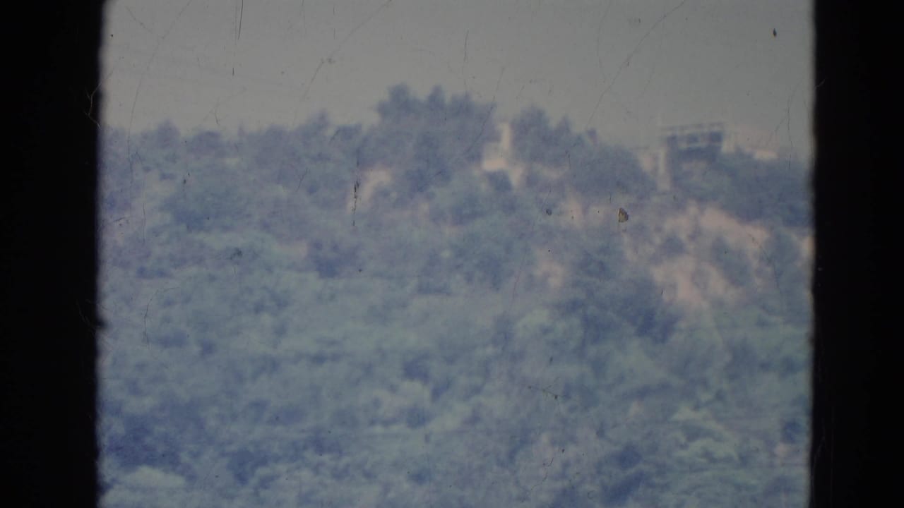 1976 Minnesota home-movie frame of a distant forested hillside and pale ridge-top shapes, divided by window framing.