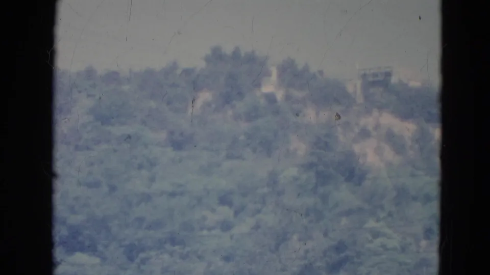 1976 Minnesota home-movie frame of a distant forested hillside and pale ridge-top shapes, divided by window framing.