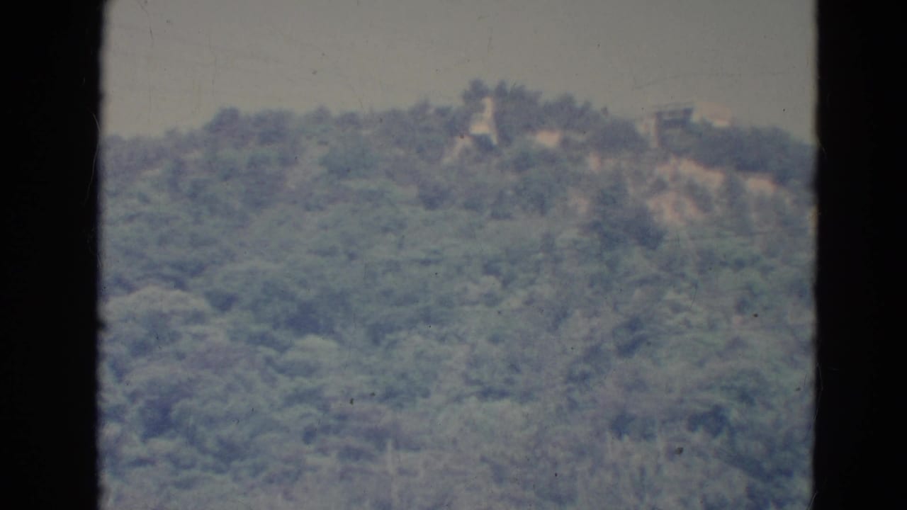 Forested hillside with faint structures near the ridge, seen through dark film-frame borders.