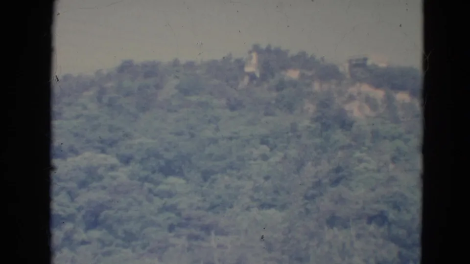 Soft 1976 Minnesota 8mm view of a forested ridge with faint clearings, distant structures, pale sky, and visible film-gate edges.