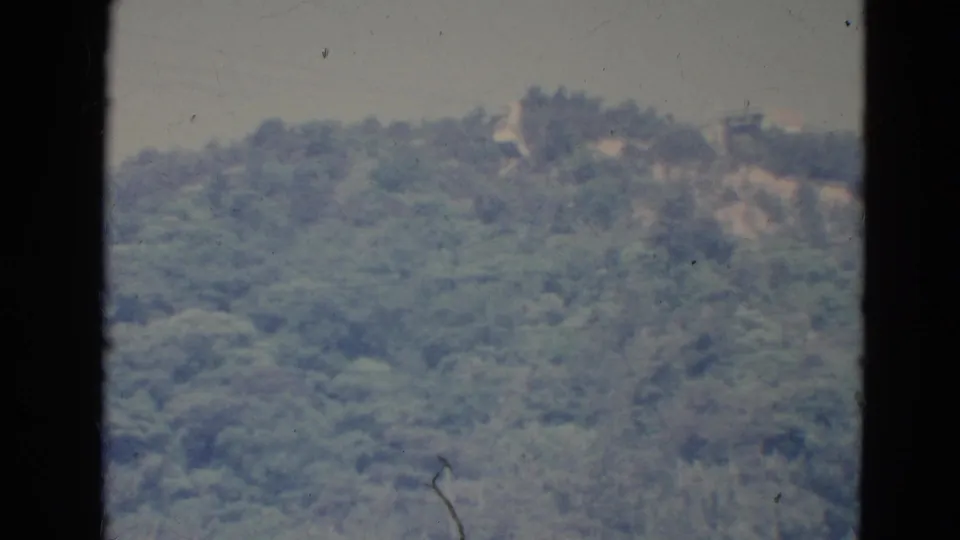 1976 Minnesota home-movie frame showing a blurred forested hillside, small ridge-top clearings, and visible 8mm surface marks.