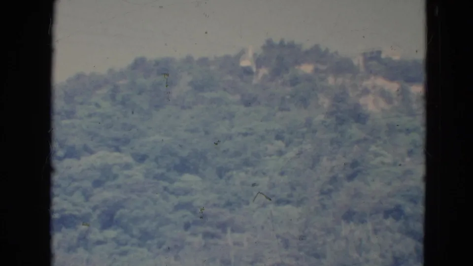 Soft 8mm frame from 1976 Minnesota showing a distant forested hillside, pale sky, faint ridge structures, and visible film dust.