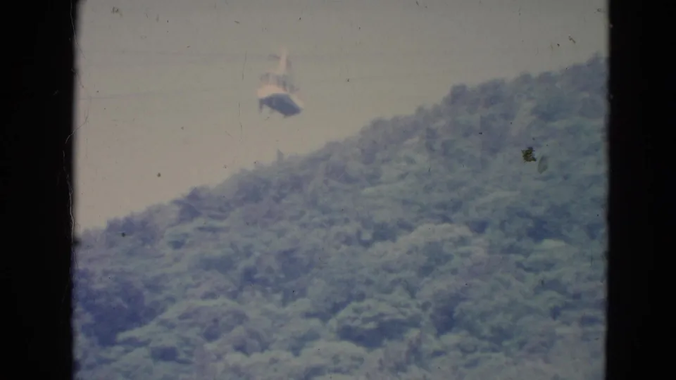 1976 Minnesota 8mm frame showing a small gondola passing over a thick green forested hillside, seen through dark frame edges.