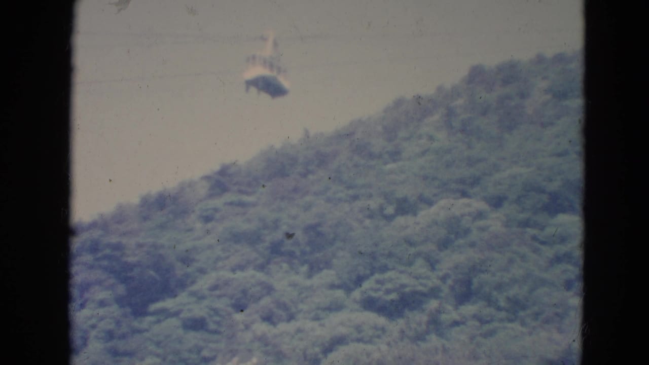 1976 Minnesota home-movie frame with a small gondola suspended over a wooded hillside, softened by faded 8mm color.