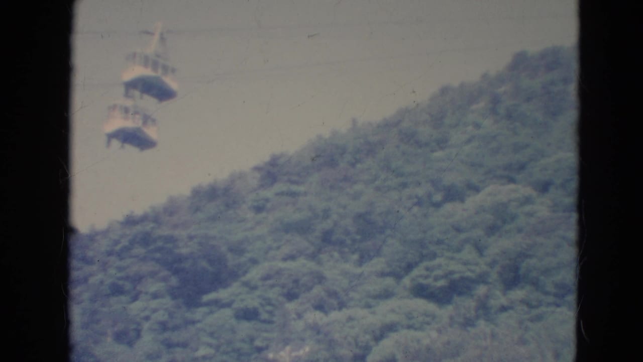 Distant 1976 Minnesota home-movie view of gondola-style cable cars crossing above dense green trees.