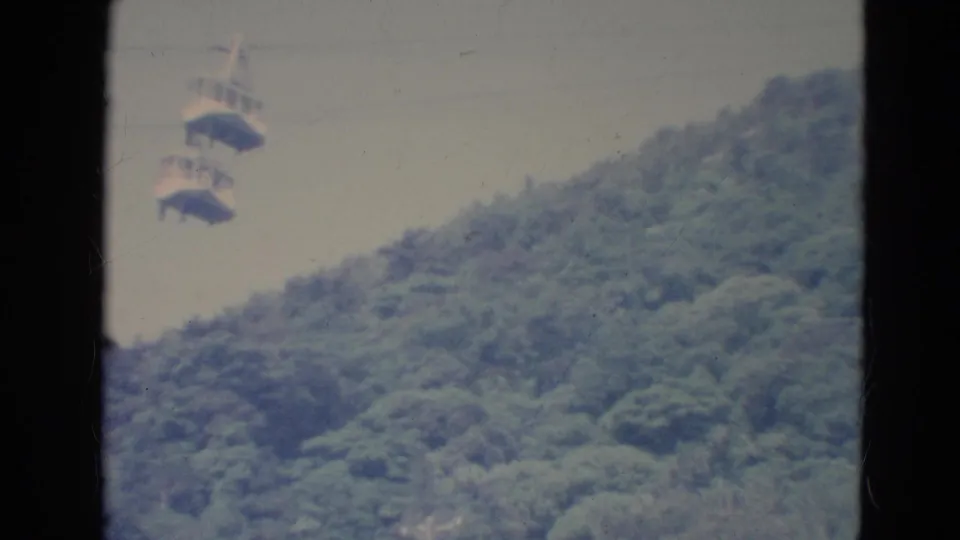 Distant 1976 Minnesota home-movie view of gondola-style cable cars crossing above dense green trees.