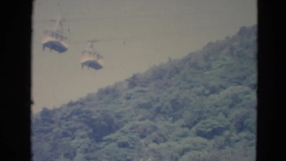 Soft 8mm frame from Minnesota in 1976 showing cable cars above a green wooded hill and pale sky.