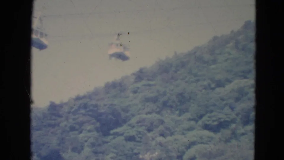 1976 Minnesota 8mm frame with a distant cable car crossing above a forested mountain slope.