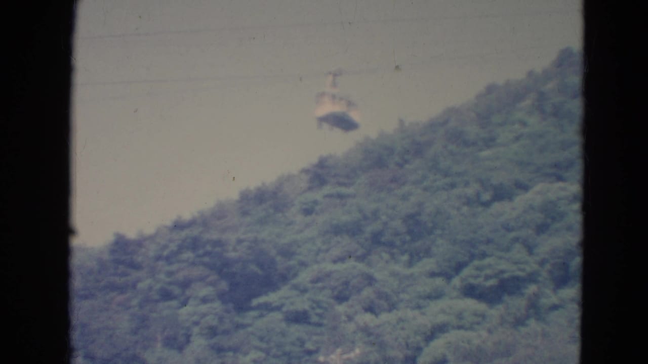 1976 Minnesota home-movie frame with a distant cable car suspended above a forested hillside, bordered by dark film or window edges.