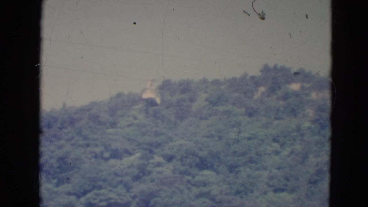 A 1976 Minnesota home-movie view of a forested ridge with faint hillside structures and visible frame borders.