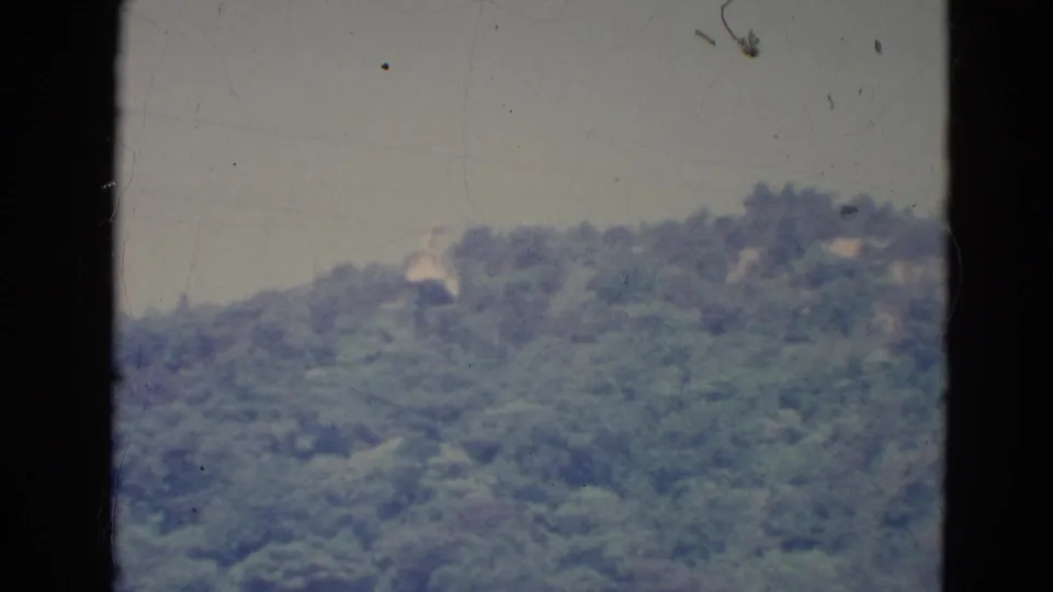 A 1976 Minnesota home-movie view of a forested ridge with faint hillside structures and visible frame borders.