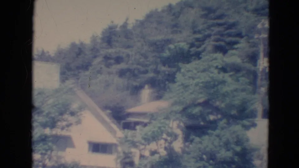 1976 Minnesota 8mm frame showing a pale house with sloped roof partly screened by trees on a wooded hillside.