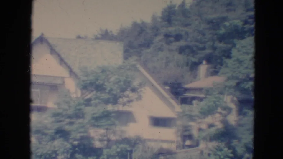 1976 Minnesota 8mm frame with rooftops, pale house walls, and dense trees beneath a wooded slope.