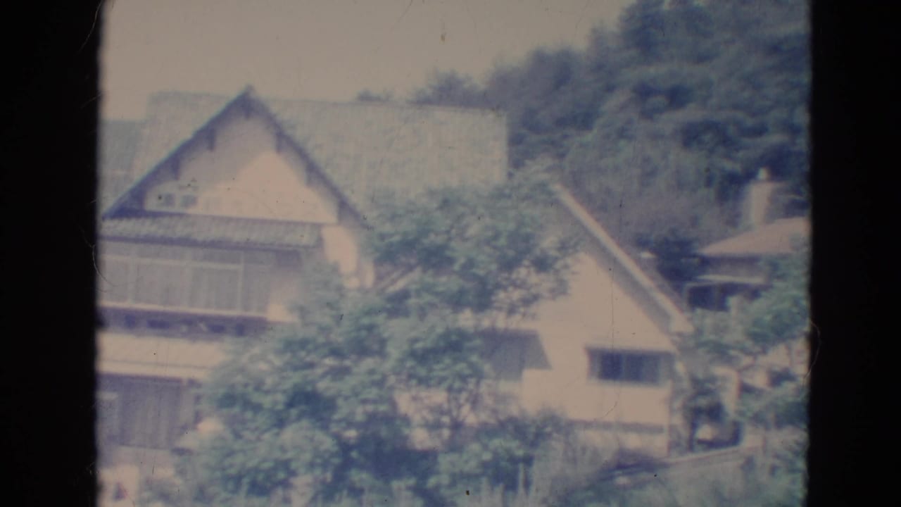 1976 Minnesota 8mm frame showing a tan house with steep roofline partly obscured by leafy trees.