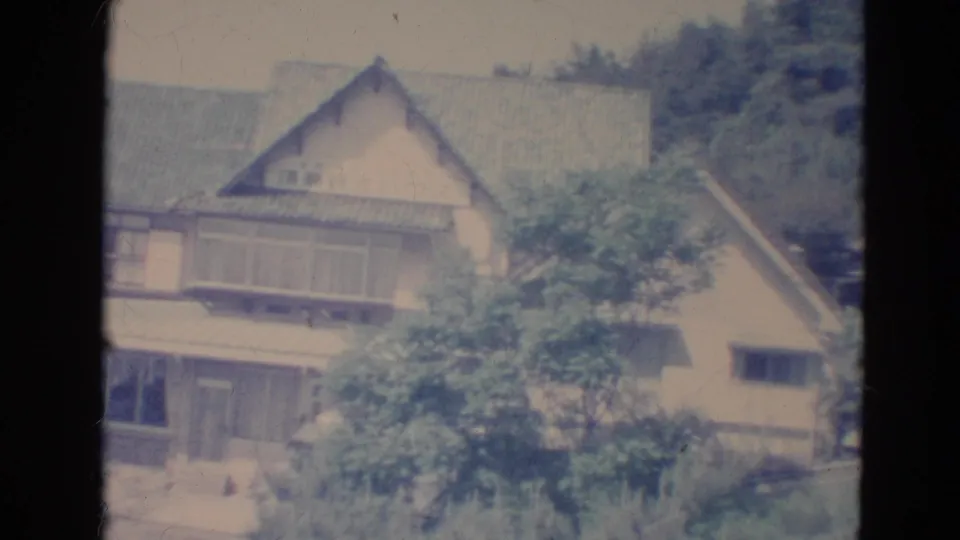 1976 Minnesota home-movie frame of a tan house and surrounding green foliage, softened by faded 8mm color.
