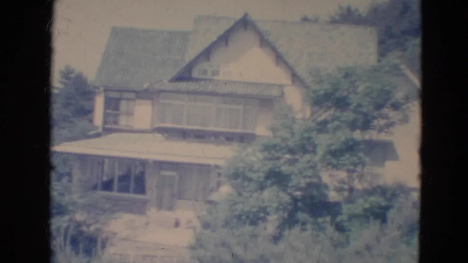 1976 Minnesota 8mm frame showing the house porch, windows, chimney, and trees from a steady outdoor viewpoint.