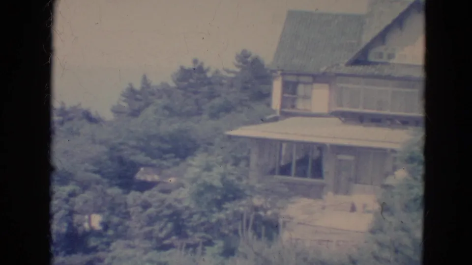 1976 Minnesota 8mm frame showing a two-story house partly obscured by dense evergreens and soft faded film color.
