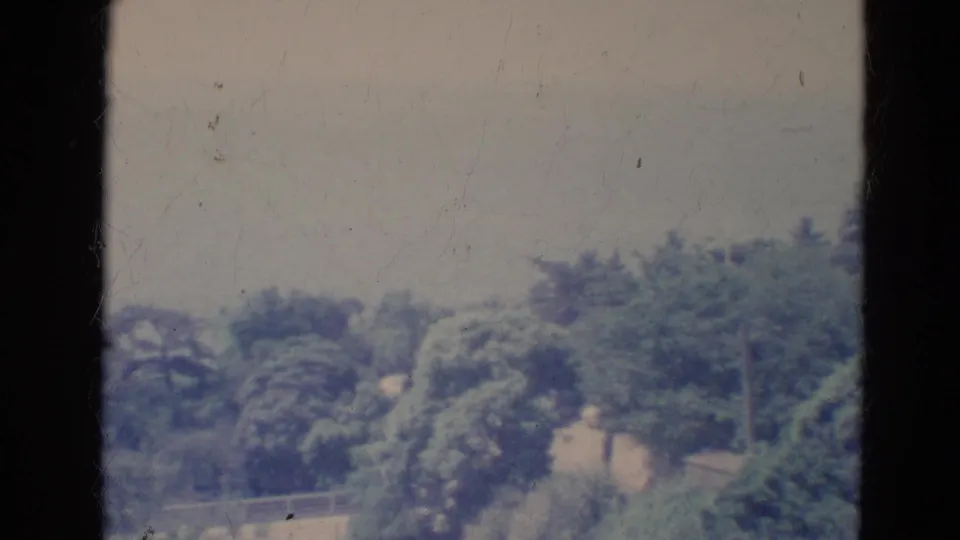 1976 Minnesota home-movie frame with trees and a railing in soft focus between dark vertical film borders.