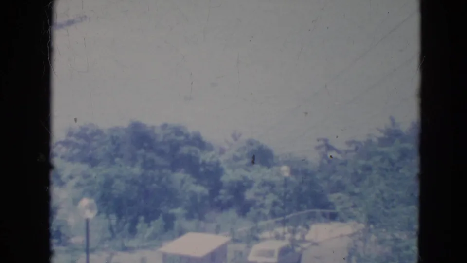 1976 Minnesota 8mm frame showing an elevated view of a tree-lined paved street with a streetlight, small shed-like structure, and parked car.