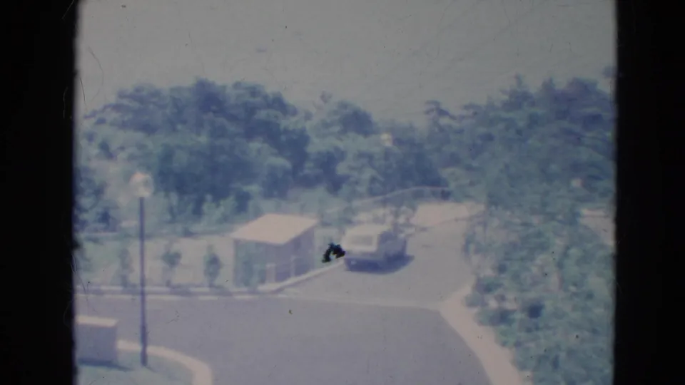 1976 Minnesota 8mm still showing a parked car beside a small structure on a paved street, framed by a streetlight, curb, and trees.