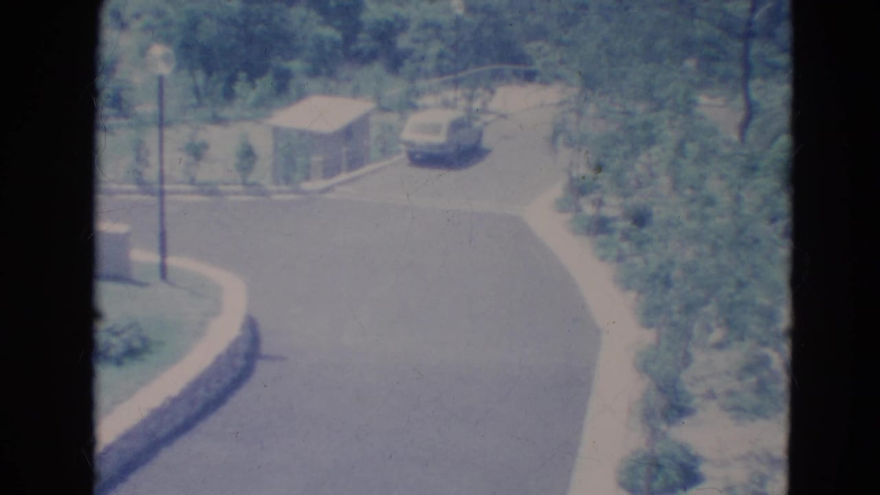 Curved suburban street with a light blue car near trees, sidewalk, shed, and streetlamp.