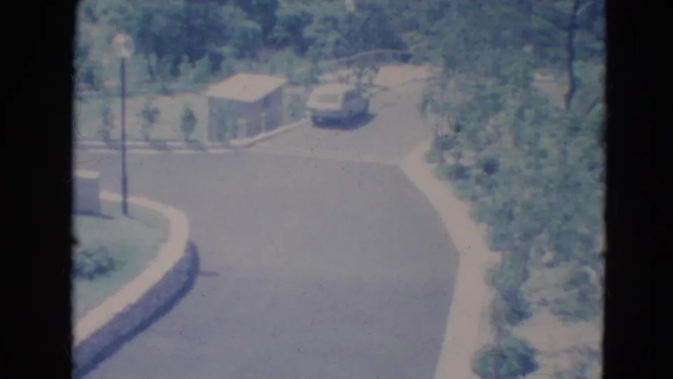 1976 Minnesota home-movie frame showing a light blue car on a curved residential street bordered by trees and sidewalk.