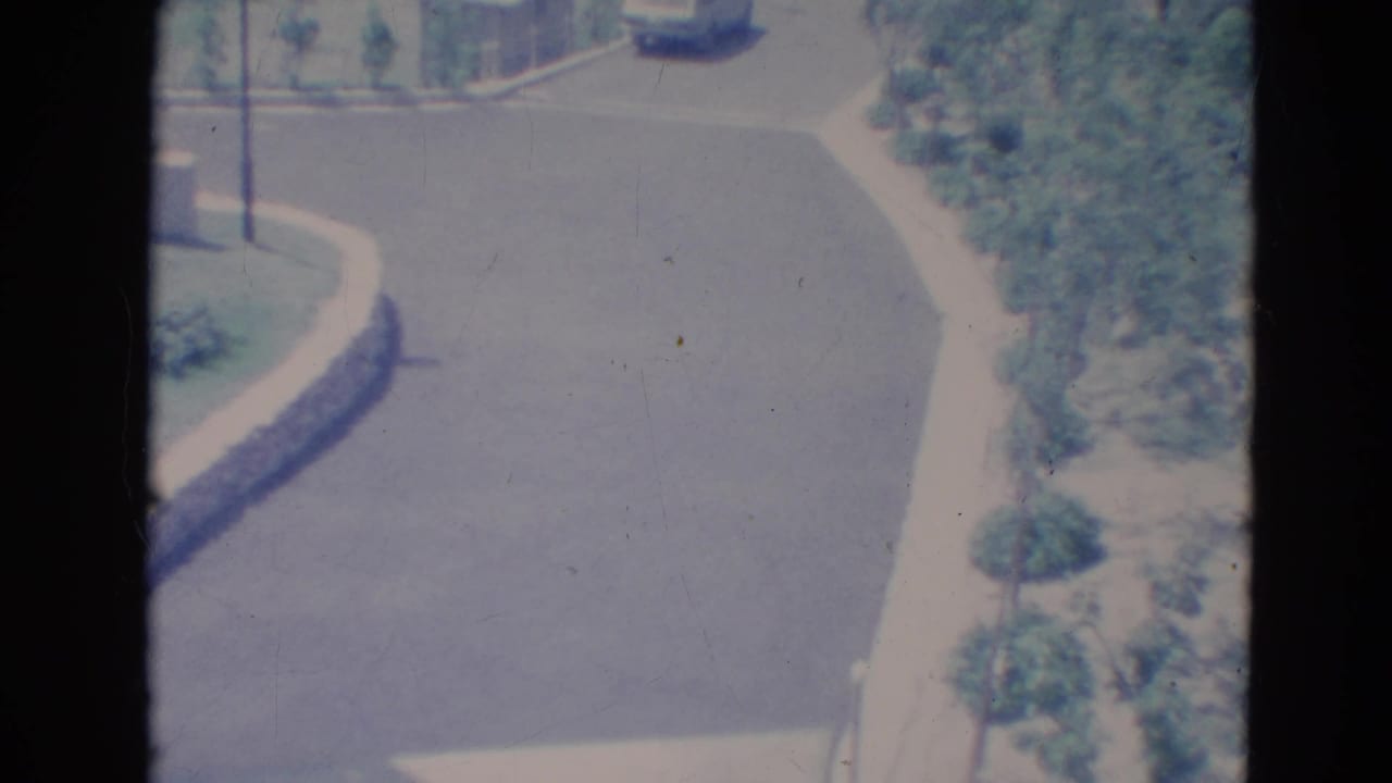 1976 Minnesota street view with a light blue car farther along a curved asphalt road bordered by greenery and sidewalk.