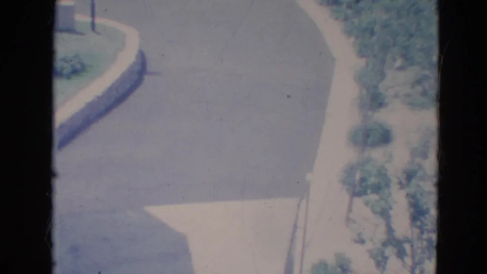 Faded 1976 Minnesota home-movie frame showing a quiet curved residential street with sidewalk, shrubs, and trees.