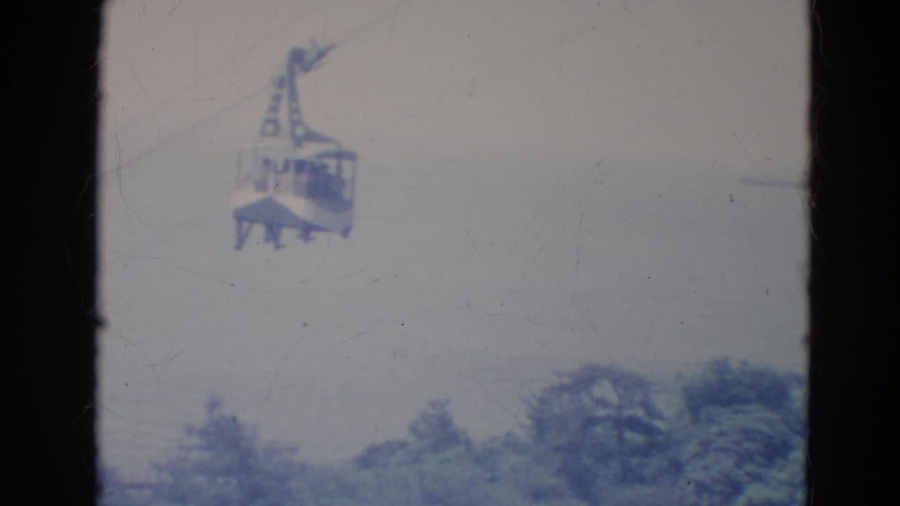 1976 Minnesota home-movie frame of a suspended skytram cabin crossing a hazy sky above trees.
