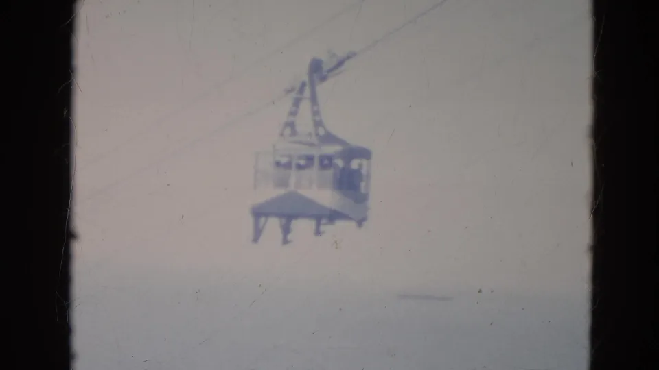 1976 Minnesota 8mm frame showing a small gondola cabin carrying passengers along overhead cables in a pale sky.