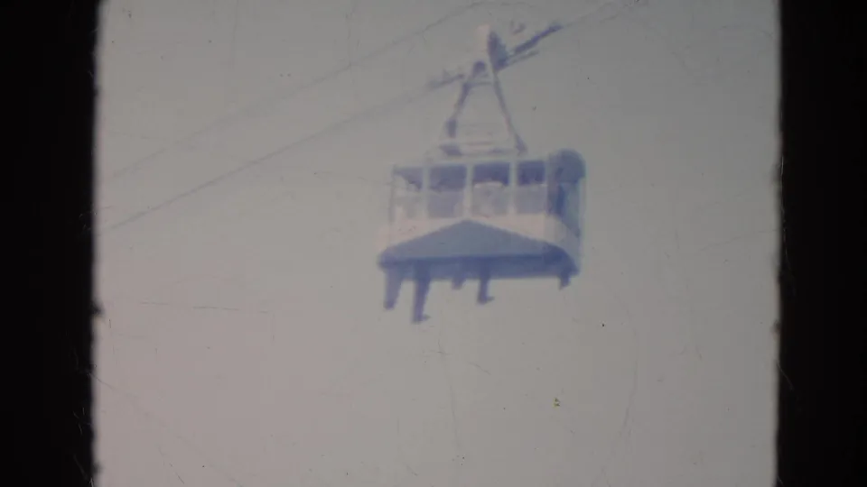 Soft 8mm frame of a Minnesota gondola cable car carrying passengers, with cables crossing the pale sky.