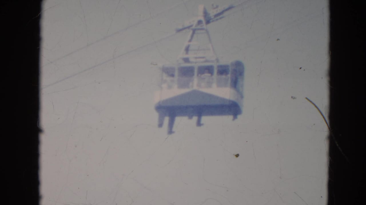 1976 Minnesota home-movie frame showing a cable car cabin on an overhead line with faint passengers visible inside.