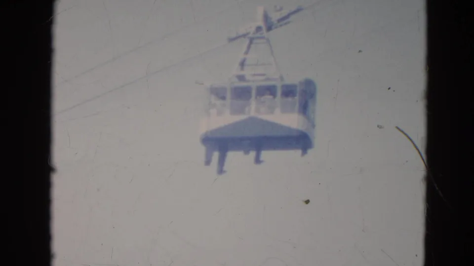 1976 Minnesota home-movie frame showing a cable car cabin on an overhead line with faint passengers visible inside.