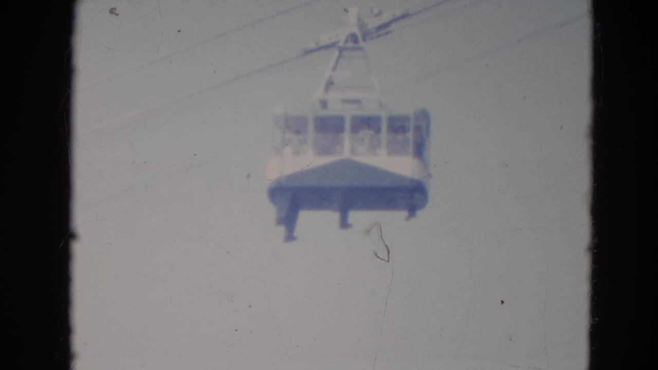 Vintage 8mm frame of a cable car suspended in an overcast Minnesota sky, with indistinct riders inside the cabin.