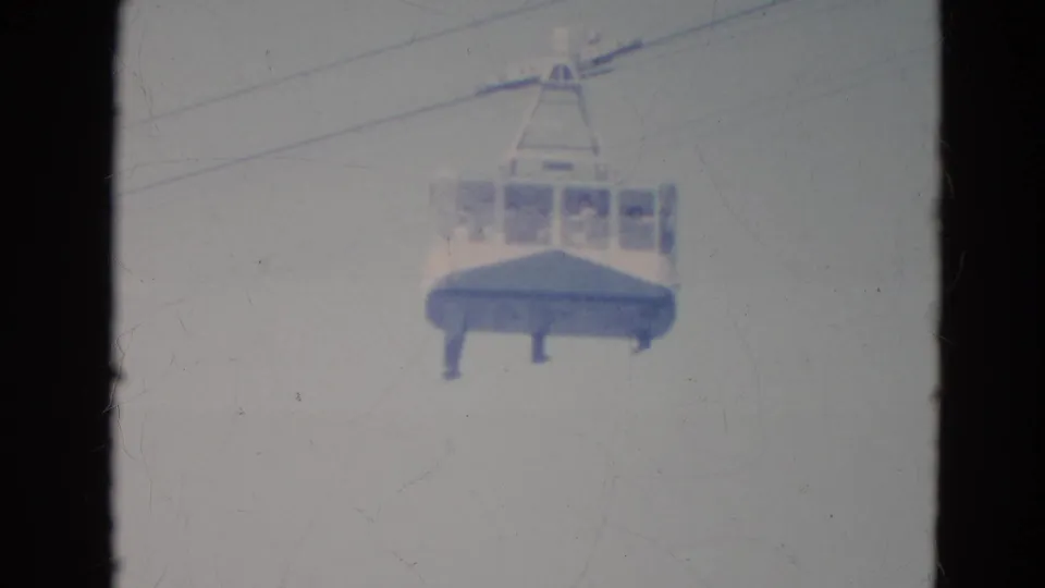 Home-movie frame showing the cable car lower in the shot, its windows and faint passengers softened by faded 8mm exposure.