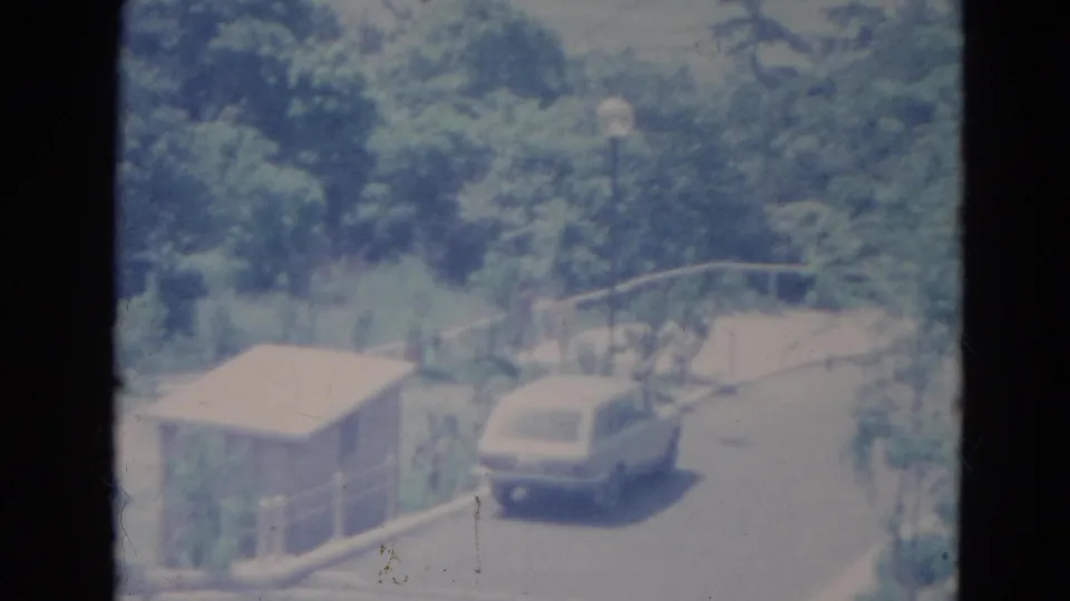 1976 Minnesota 8mm frame showing a pale station wagon near a roadside shed, guardrail, lamppost and dense summer trees.