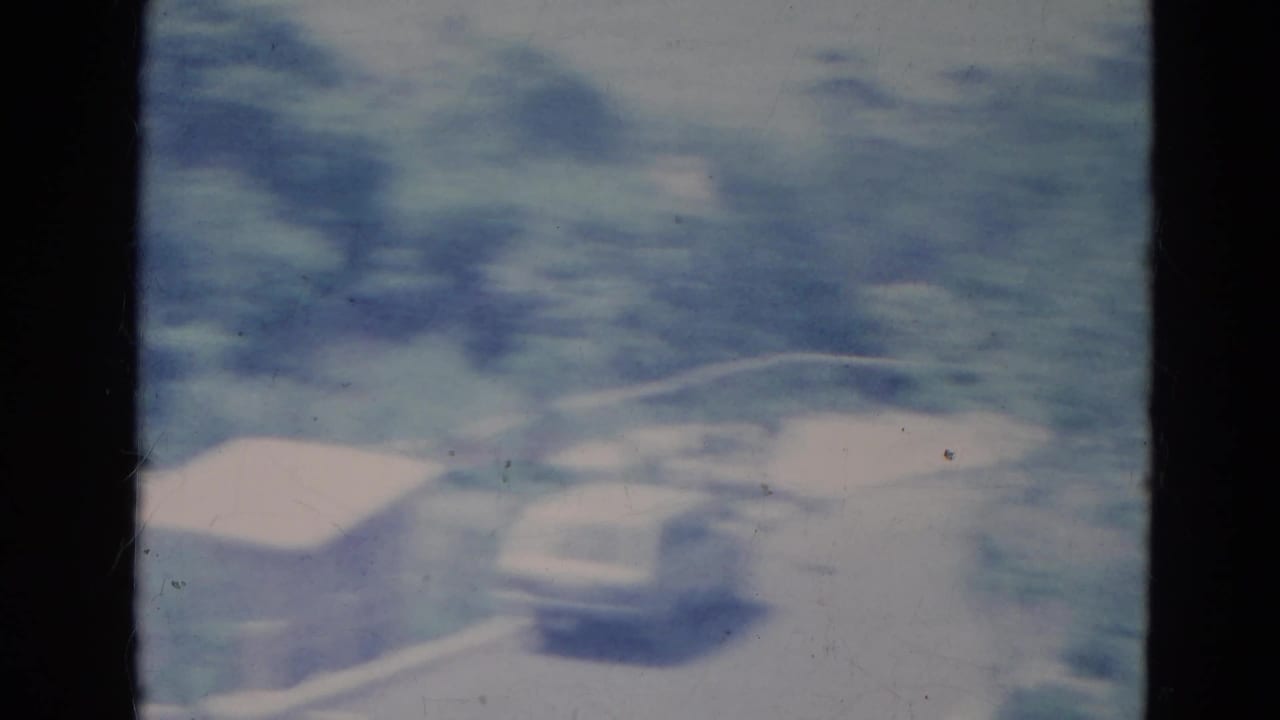 Motion-blurred 1976 Minnesota 8mm frame with a pale station wagon, small shed and leafy roadside area.