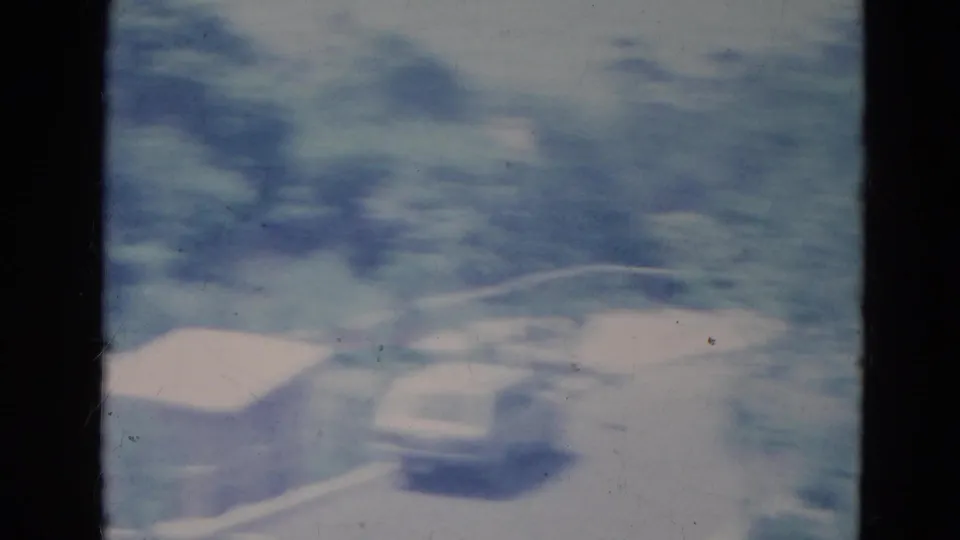 Motion-blurred 1976 Minnesota 8mm frame with a pale station wagon, small shed and leafy roadside area.