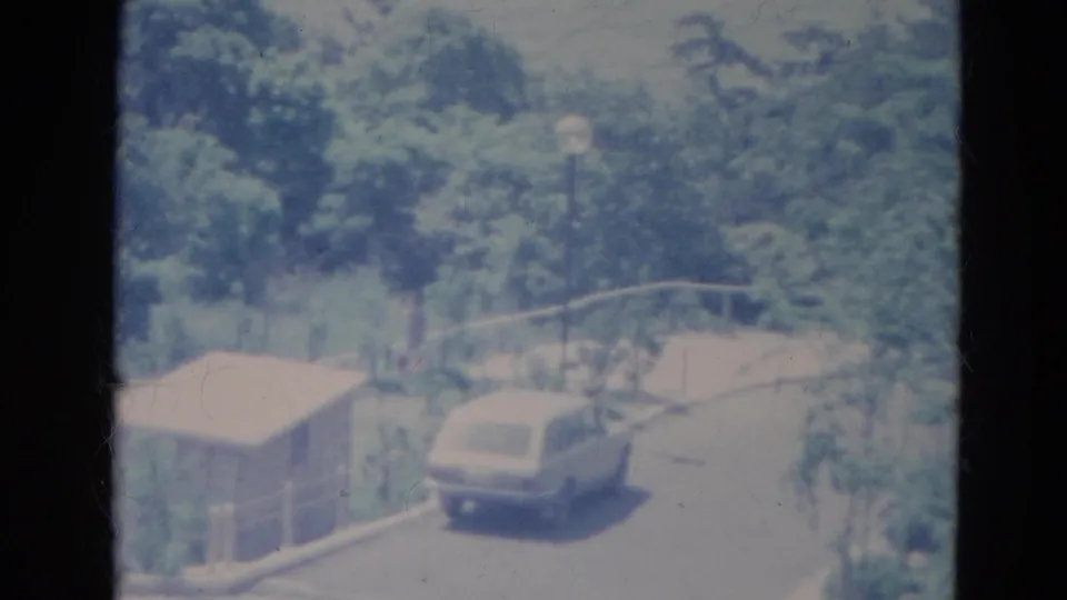 1976 Minnesota home-movie frame showing a light station wagon by a shed, guardrail and tree-lined road in bright light.