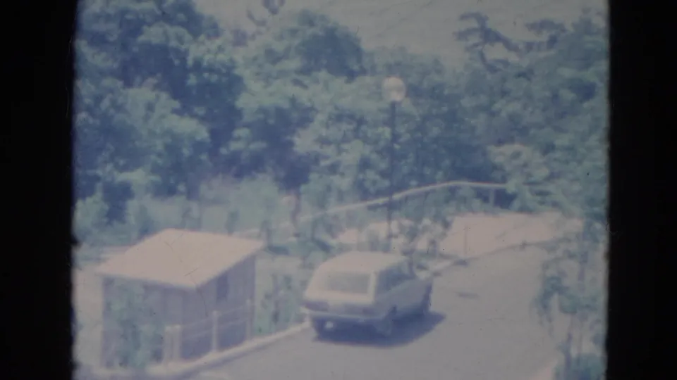 1976 Minnesota 8mm frame with a pale station wagon on a leafy road near a small shed, guardrail and globe lamppost.