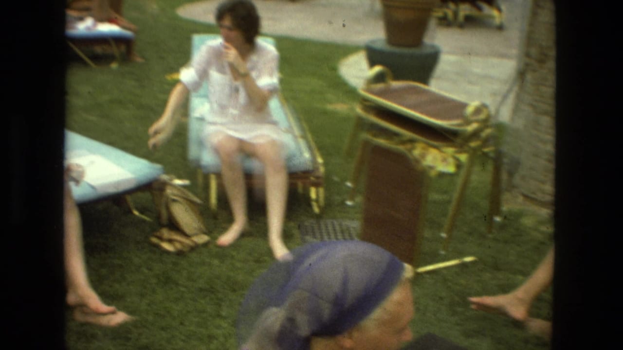 1978 Las Vegas frame with a seated woman in a pale outfit on a blue lounge chair, surrounded by grass, bags, and outdoor chairs.