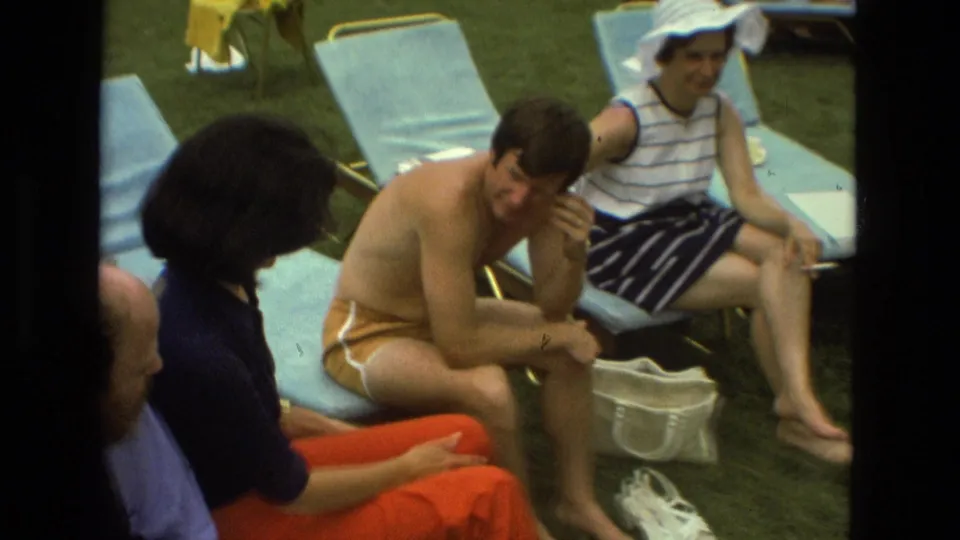 1978 Las Vegas frame with adults seated closely on blue lounge chairs, a man in orange shorts, and a woman in a striped dress.