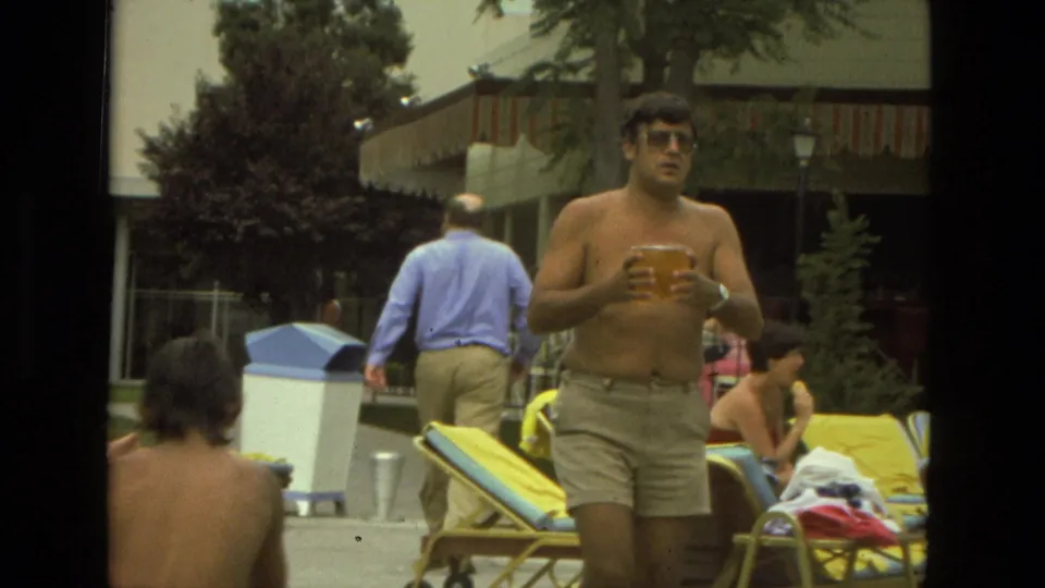 1978 Las Vegas home-movie frame of a shirtless adult man walking with a plastic cup near lounge chairs and other people outdoors.