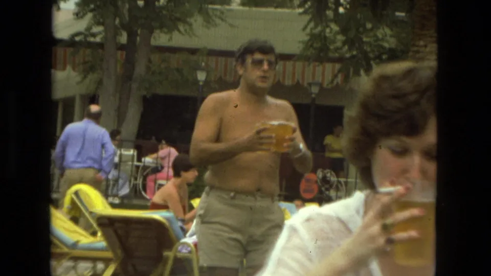 1978 Las Vegas outdoor leisure frame with a man holding a plastic cup, another person drinking in the foreground, and lounge chairs behind them.