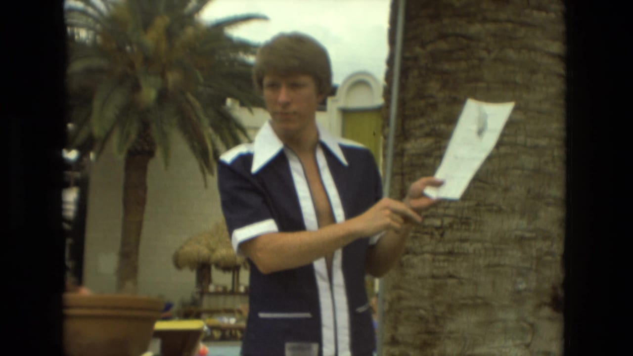 Young man in a blue and white 1970s shirt holds papers near palm trees at a Las Vegas resort area.