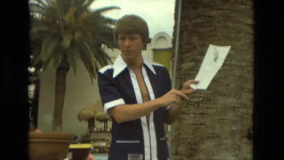 Young man in a blue and white 1970s shirt holds papers near palm trees at a Las Vegas resort area.